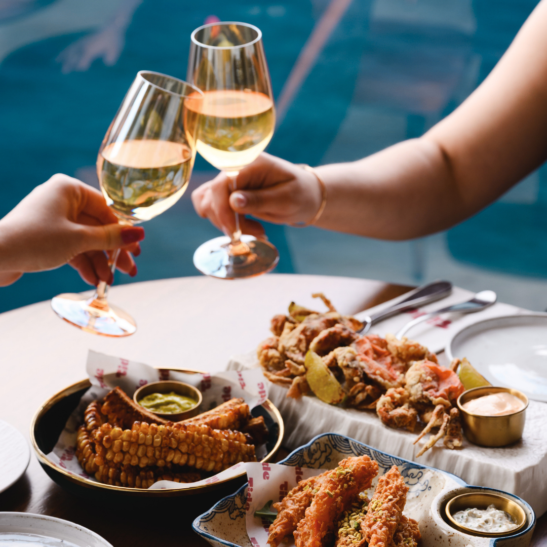 Two people clink wine glasses over a table piled with fried appetizers like corn on the cob and fried shrimp.