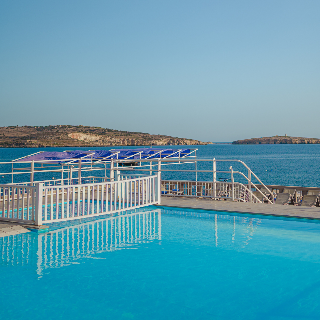 Outdoor pool by the sea with white railings and blue canopies on a sunny day, island in the distance
