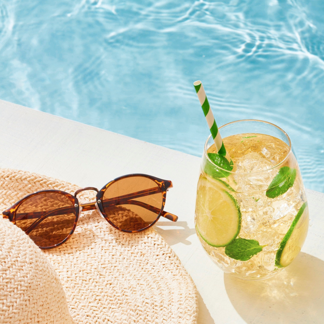 Poolside scene with brown sunglasses resting on a woven straw hat beside a glass of lemon-mint iced drink by the pool edge.